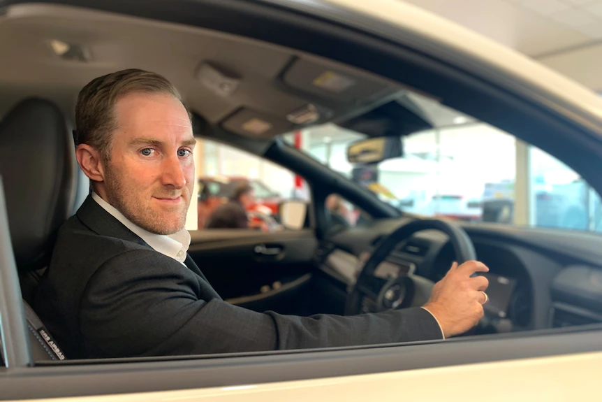 A Nissan employee sits in a parked car holding the steering wheel and looking at the camera