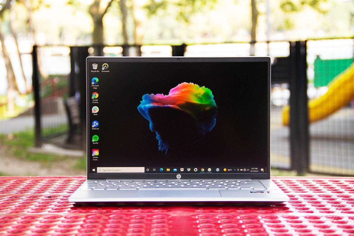 The HP Pavilion Aero 13 open on a red picnic table with a playground in the background. The screen displays a black desktop background with a multicolored cloud in the center.